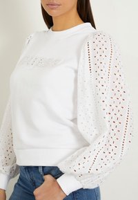 Sudadera blanca con el logo "GUESS" bordado, con mangas abullonadas de encaje texturizado y puños acanalados. Corte casual, diseñada para la comodidad.