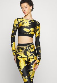 Crtkana crna i zlatna kratka majica dugih rukava s odgovarajućim tajicama, koja sadrži detalj od lanca i uski dizajn.