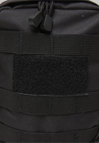 Primer plano de un bolso táctico negro con tela texturizada, correas de cincha, un parche rectangular de Velcro y dos cierres de cordón con cordón ajustable.