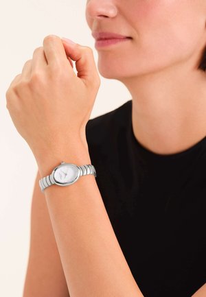 Silberne Armbanduhr mit rundem Zifferblatt und weißem Ziffernblatt, mit einem strukturierten Metallarmband. Getragen am Handgelenk mit neutralem Hautton.