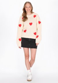 Cremfarbener Strickpullover mit verstreuten roten Herzmustern, kombiniert mit einem schwarzen Denimrock. Das Modell trägt weiße Socken mit Herzen und Sneakers.