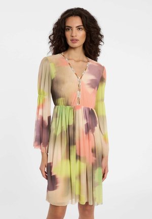 Frau mit lockigem Haar, die ein mehrfarbiges knieumspielendes Kleid mit langen Ärmeln und V-Ausschnitt trägt, der mit einem gemusterten Rand verziert ist.