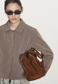 Brown suede bucket bag with a textured finish and light-colored piping. Model wearing a gray jacket with buttons, showcasing a casual look.