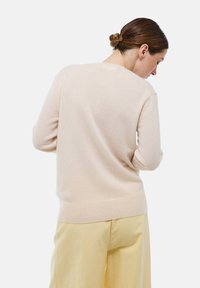 Femme aux cheveux bruns en chignon, portant un pull beige clair et un pantalon jaune pâle, tournée légèrement sur le côté contre un fond blanc.