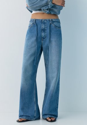 Osoba w niebieskich dżinsach z szerokimi nogawkami i wysokim stanem, z deninowym paskiem ze sznurkiem oraz czarnych sandałach z odkrytymi palcami na jednolitym jasnym tle.