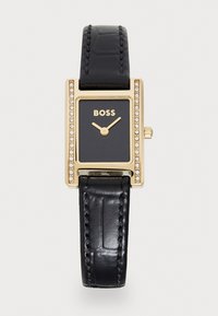 Relógio retangular dourado com mostrador preto. A bracelete de couro preto apresenta uma textura de crocodilo. Acentuado com pedras em torno da luneta.