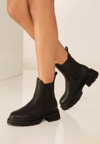 Botas de tobillo de cuero negro con paneles laterales elásticos, punteras redondas y suelas de goma negra texturizada, que cuentan con un tacón bajo en bloque.