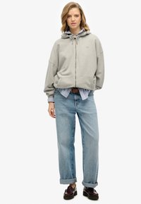 Sudadera con cremallera de color gris claro, con capucha ajustable, bolsillos frontales, sobrecamiseta de rayas azules, combinada con jeans de mezclilla claros y mocasines oscuros.