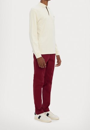 Man draagt een crèmekleurige poloshirt met lange mouwen, bordeauxrode broek en witte sneakers met zwarte strepen, staand tegen een effen achtergrond.