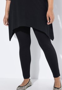 Zwarte leggings van rekbaar materiaal, een aansluitend ontwerp en een gladde textuur. Gecombineerd met een zwarte tuniek top en gepatternde platte schoenen.
