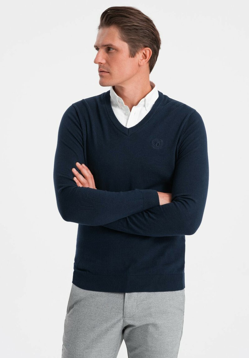 Maglione blu navy a V in morbido tessuto a maglia, con una vestibilità slim e orlo a coste. Indossato sopra una camicia bianca con colletto, senza motivi prominenti.