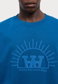 Blue cotton t-shirt featuring a graphic print in a darker blue, depicting a sun and letters "WW" with the text "DOUBLE A BY WOOD WOOD."