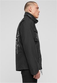 Černá bunda s vysokým límcem, několika předními kapsami a složitým grafickým vzorem na zádech. Hladká textura a volný střih.