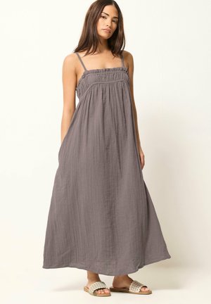 Junge Frau trägt ein lockeres, graues, knöchellanges ärmelloses Kleid mit dünnen Trägern und beigen Slip-on-Sandalen, steht vor einem schlichten Hintergrund.