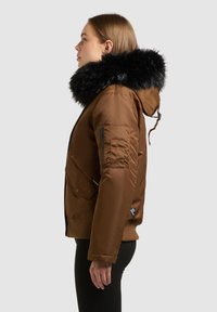 Hnědá prošívaná bunda s černou kapucí z umělé kožešiny. Obsahuje kapsu na zip na rukávu a přední kapsy s klopami, hladkou texturu s žebrovaným lemem.