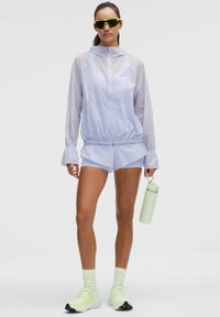 Rompevientos de color lila claro con cierre frontal, puños elásticos y pantalones cortos a juego. Combinado con gafas de sol tintadas en verde y zapatillas deportivas de color verde claro.