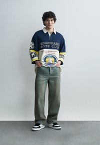 Ung mann står med hendene i lommene, iført en overdimensjonert marineblå og hvit Billionaire Boys Club rugbyskjorte, falmede vide jeans og joggesko.