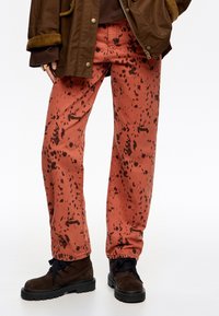 Rudos džinsai su juodu purslų raštu, turintys laisvą siluetą. Sušalinta su tamsiais batais ir rudais švarkais.