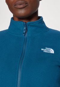 Chaqueta de forro polar azul con cuello alto y cierre de cremallera. Presenta un logo blanco en la parte superior izquierda. Textura suave, diseño informal.