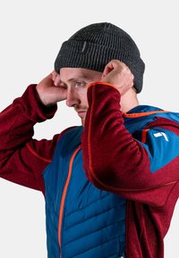 Dark gray knit beanie, ribbed texture, with a tag. Worn with a red fleece jacket and blue insulated vest; orange accents on the zipper.