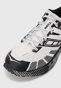 Baskets de course en noir et blanc avec un matériau en mesh, des lacets larges, une semelle texturée et des motifs géométriques sur le côté. Modèle : Speedgoat 2.