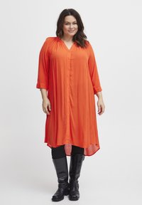 Narancssárga, gombos ruha 3/4-es ujjakkal, V-kivágással és ívelt aljjal. Fekete, testhezálló leggings-szel és térd fölé érő csizmával párosítva. Sima textúra.