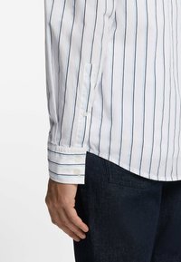 Wit overhemd met blauwe verticale strepen, knoopmanchetten en een lichte glans. Gecombineerd met donkere denimbroek met een zakdetail.