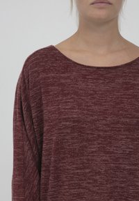 Burgundská oversized blůza s kulatým výstřihem, která má strukturovanou tkaninu a volný střih. Materiál vypadá měkký s jemnými variacemi v barvě.