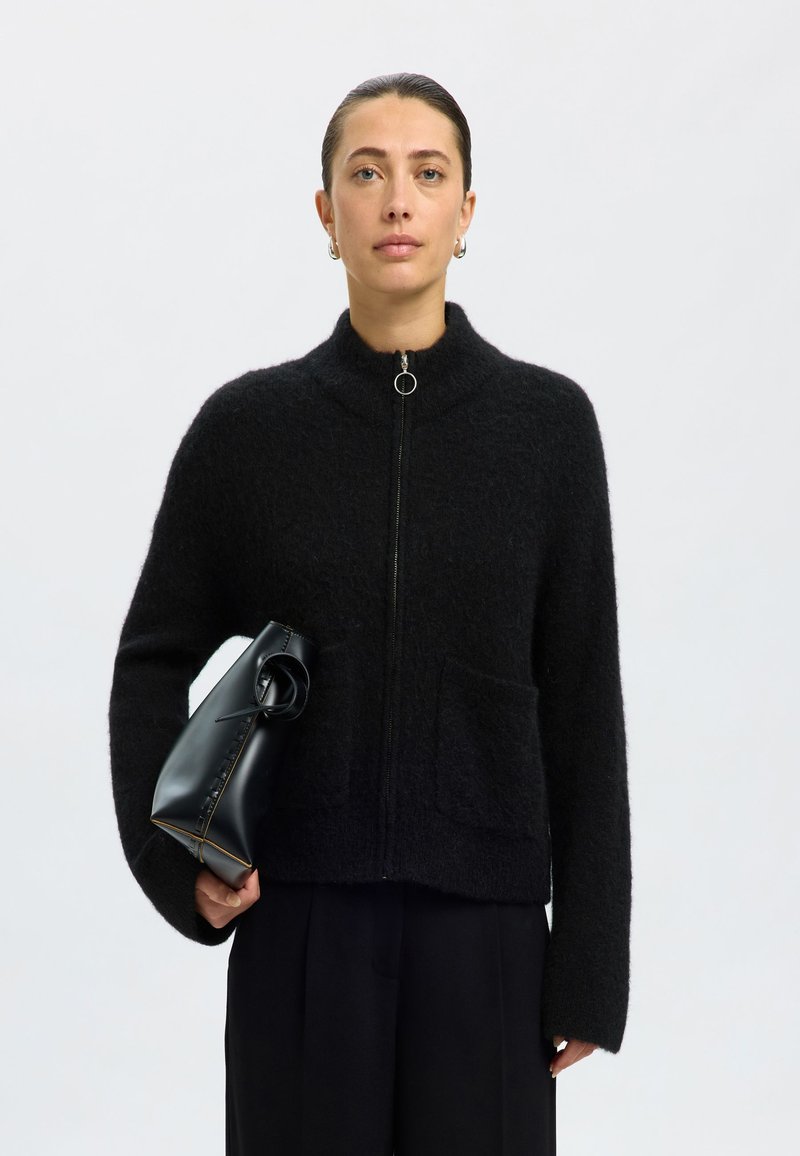 Crna vuna zip-dukserica s visokim ovratnikom, plišastog izgleda, s prednjim džepom, u kombinaciji sa strukturiranom crnom kožnom torbom.