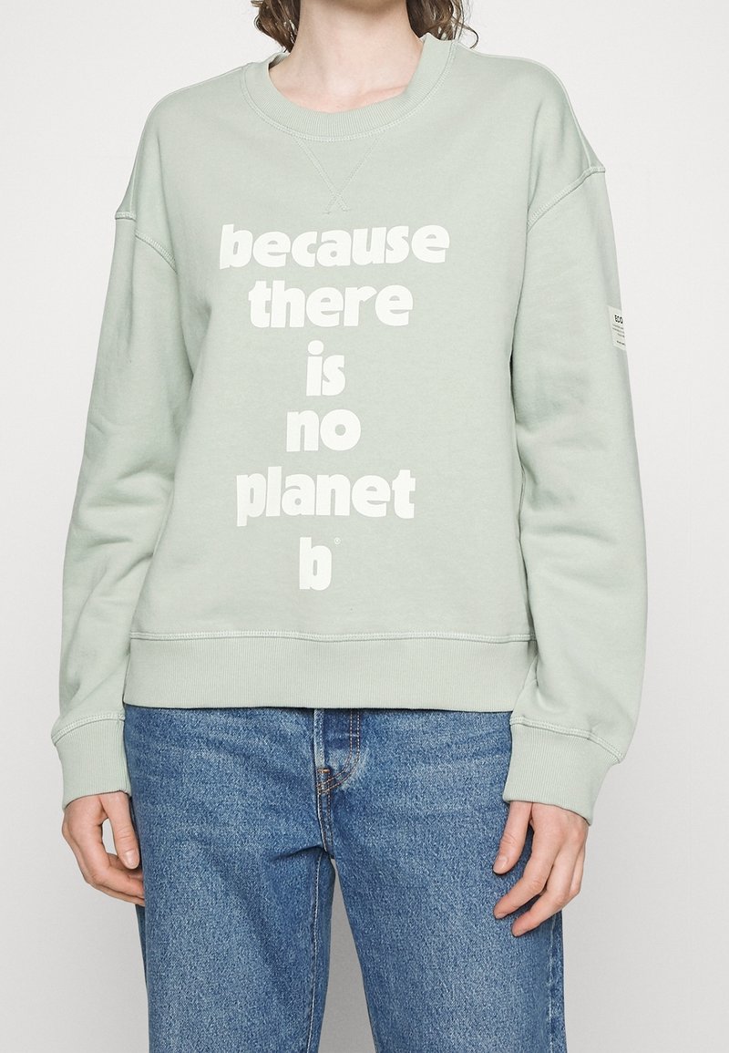 Personne portant un sweat-shirt vert clair avec un texte blanc « because there is no planet b » et un jean bleu sur un fond uni.