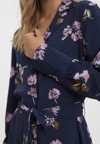 Detailní záběr osoby v námořnických šatech s růžovým květinovým potiskem a páskem v oblasti pasu, ruka blízko límce, ukazující dlouhé rukávy a knoflíkovou úpravu.