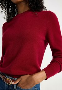 Rød strikket sweater med rund halsudskæring, lange ærmer og ribbede manchetter. Glat tekstur, stylet med denimjeans og et brunt bælte.