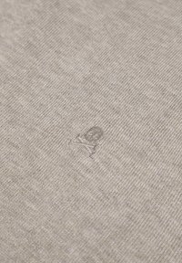 Material tricotat gri, cu un mic logo brodat de craniu, de culoare gri deschis. Textura este moale și ușor melanjată.