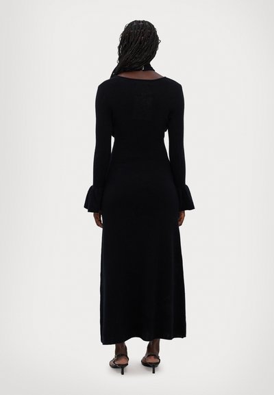 Robe maxi noire à manches longues avec un corsage ajusté, des poignets évasés et une texture lisse. Le dos présente un design ouvert. Style simple et élégant.