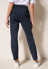 Joggers navy avec taille élastiquée, deux poches arrière et jambes fuselées, associés à des baskets blanches sur un fond neutre.