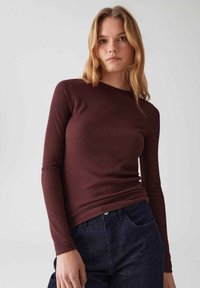 Giovane donna con capelli castani chiari che indossa un top a maniche lunghe, a costine, di un intenso borgogna scuro e jeans blu scuro, su uno sfondo grigio neutro.