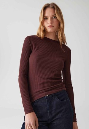 Jonge vrouw met lichtbruin haar, gekleed in een lange mouw, geribbelde, donkerbordeaux top en donkerblauwe jeans, tegen een effen grijze achtergrond.