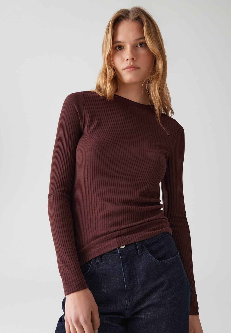 Giovane donna con capelli castani chiari che indossa un top a maniche lunghe, a costine, di un intenso borgogna scuro e jeans blu scuro, su uno sfondo grigio neutro.