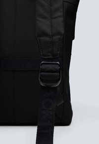 Mochila negra con una asa de mango texturizado y una hebilla de plástico prominente. La correa está grabada con el nombre de la marca.