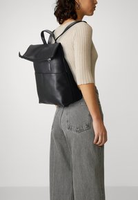 Svart lær ryggsekk med klaff, glidelåslukking, frontlomme og justerbare stropper; sammen med grå høydeformat jeans.