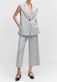 Žena v světlém šedém bezrukávovém blejzru a odpovídajících širokých kalhotách do půli lýtek, s černými block-heeled Mary Jane botami.