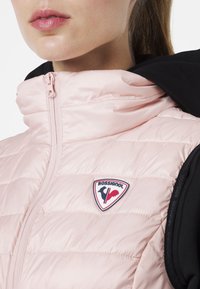 Svetle růžová nafouklá vesta s vysokým límcem, předním zipem a černou kapucí. V oblasti levého prsu se nachází nášivka loga Rossignol.