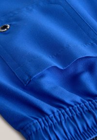 Tissu bleu avec une poche à fermeture pression, un ourlet élastique froncé et une texture lisse. Détail de couture visible.