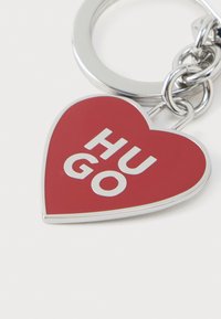 Porte-clés en forme de cœur rouge avec une ferrure argentée, présentant le mot "HUGO" en lettres blanches et en gras. Finition lisse et brillante.