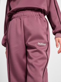 Barn iført lilla Hummel Performance joggingbukser og matchende sweatshirt, der viser elastisk talje og sidelommer.