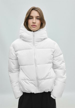 Femme aux cheveux châtain clair portant une veste matelassée blanche avec un col montant et les mains dans les poches, sur un fond clair uni.