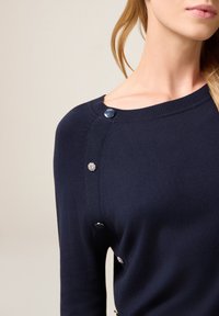 Granatowy sweter z dzianiny z dekoltem typu boat neck, dekoracyjnymi srebrnymi guzikami na ramionach oraz gładką fakturą.
