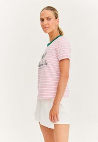 Kobieta z blond włosami w kucyku, ubrana w różowo-białą koszulkę w paski i białe szorty, stojąca bokiem na jednolitym tle.