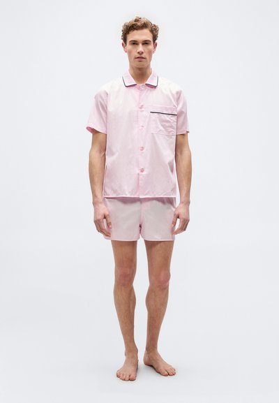 Νεαρός άνδρας στέκεται ξυπόλητος φορώντας ανοιχτό ροζ πουκάμισο με κοντό μανίκι και κουμπιά και αντίστοιχα σόρτς, μπροστά σε απλό λευκό φόντο.