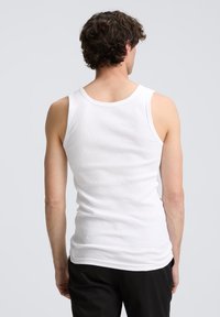Débardeur en coton blanc avec une texture côtelée. Il présente un col rond et des emmanchures basses, porté avec un pantalon noir. Vue arrière montrée.